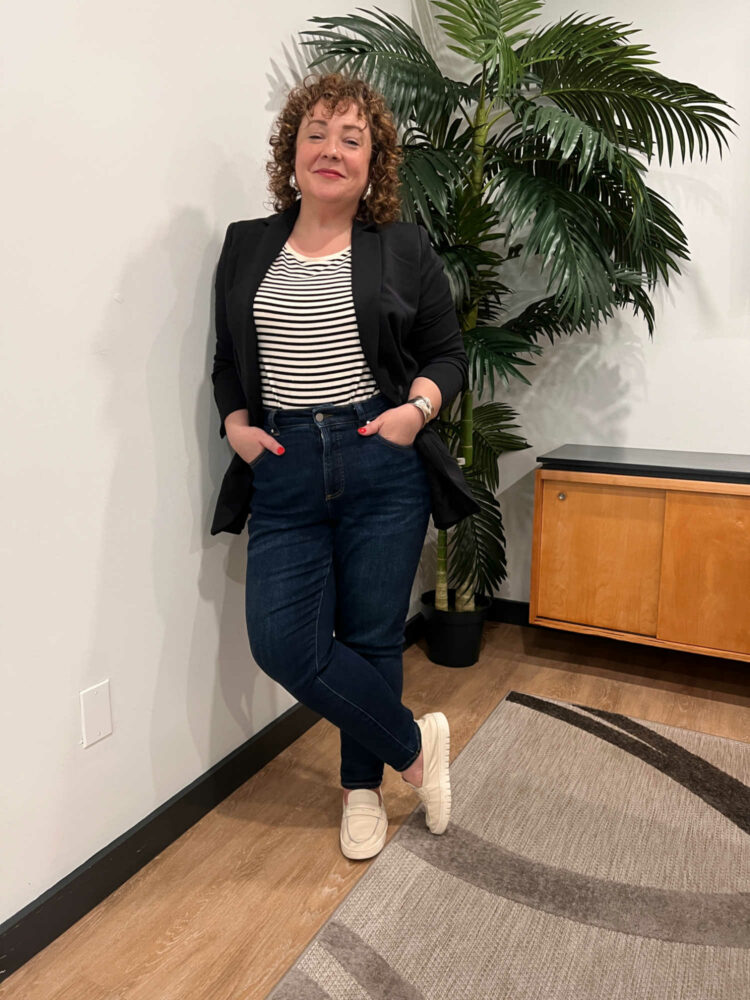 Alison leaning against a wall, wearing the Universal Standard All Day blazer in black over a cream and white stripe Tee Rex tucked into dark Joni jeans, On her feet are ivory Vionic loafers