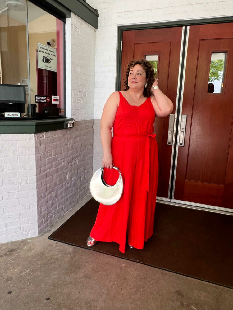 Alison in the Universal Standard linen Promenade jumpsuit in orange, holding a silver chrome Moon bag from STAUD and wearing silver platform heeled sandals from Aerosoles
