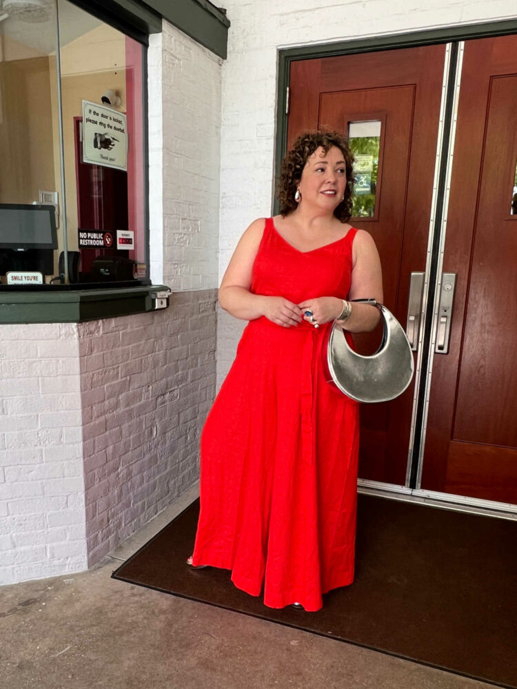 Alison holding the STAUD chrome Moon bag while standing in front of a movie theater, wearing an orange linen wide leg jumpsuit