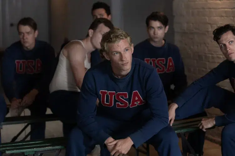 The character Joe Rantz from the movie The Boys in the Boat sitting on a bench in a USA sweater looking up