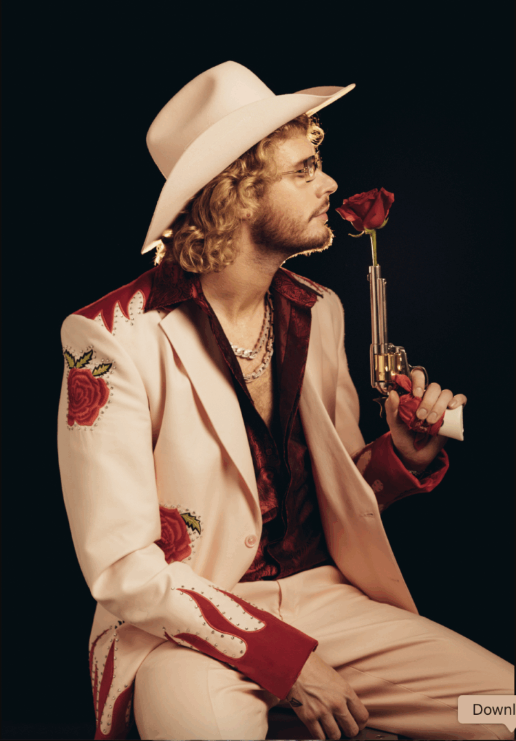 Yung Gravy in a white suit and cowboy hat holding a gun with a rose stuck in the barrel and he's smelling the rose
