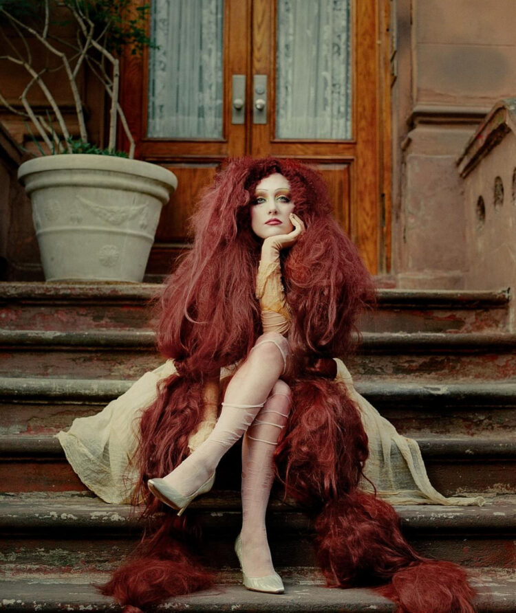 chappell roan in a very long red wig sitting on the steps of a new york city brownstone
