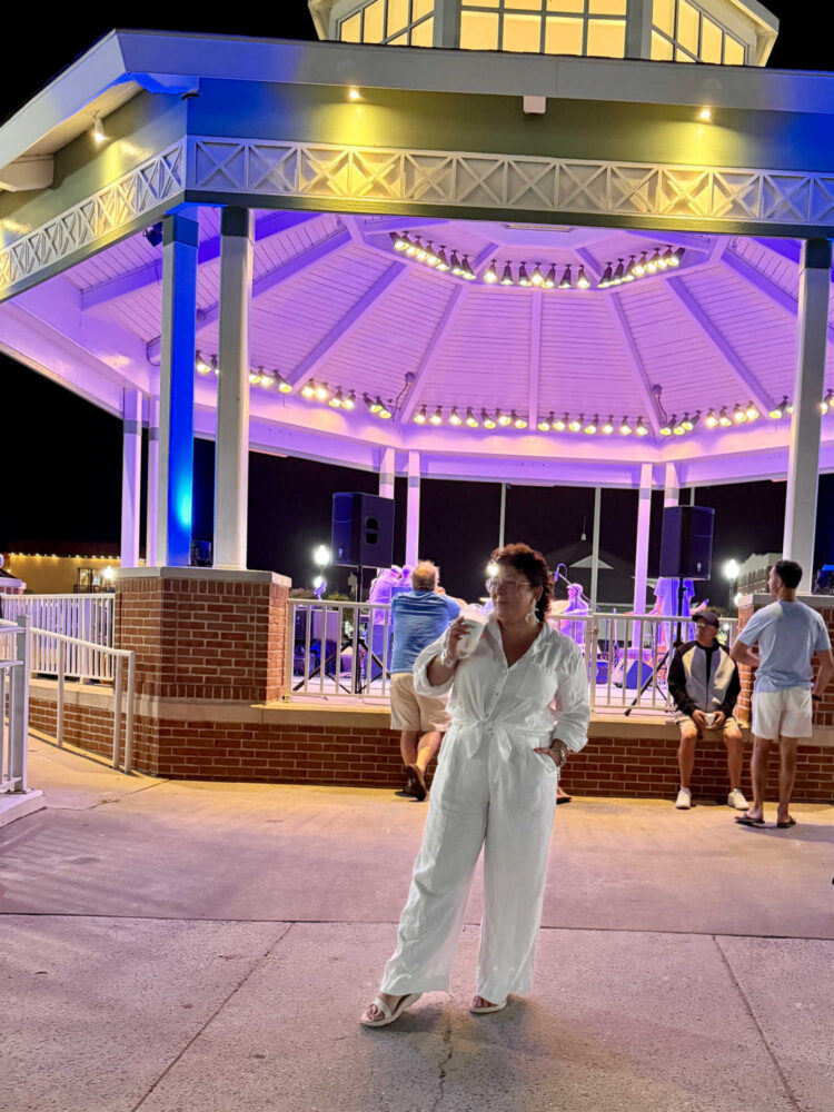 Alison in a white linen shirt tied at the waist and white linen pants standing in front of the Rehoboth Beach bandstand