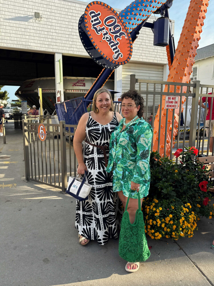 Debbie and Alison at Funland in Rehoboth Beach, August 2025. Debbie is in a black and white print linen mxi dress and Alison is in a green palm print shirt and matching shorts, holding a green macrame tote