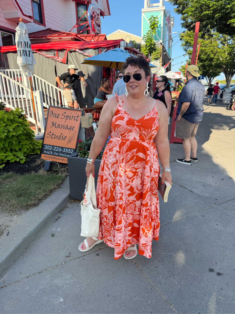 Alison of Wardrobe Oxygen in a coral and white print Land's End maxi length cotton voile sundress, holding a canvas tote and a book