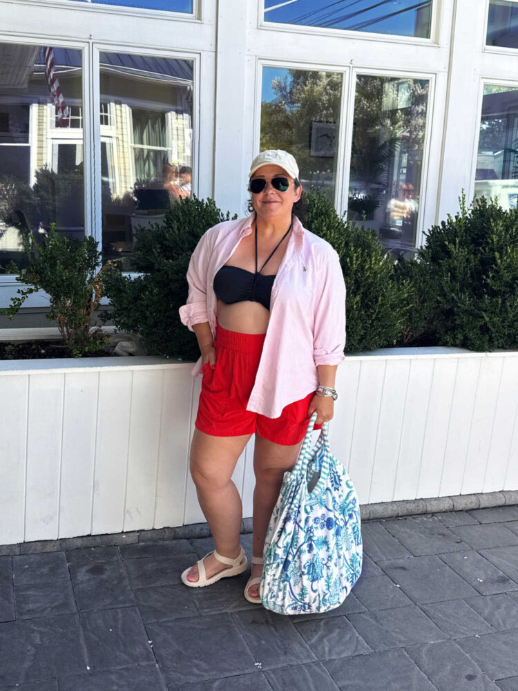 Alison Gary of Wardrobe Oxygen in a black bikini with a pink oxford shirt open and red athletic shorts. She has a blue floral cotton tote in her hand.