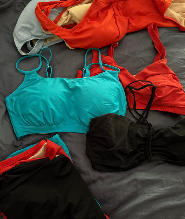 two-piece and one-piece swimsuits in turquoise, blue, black, and orange lying on a bed