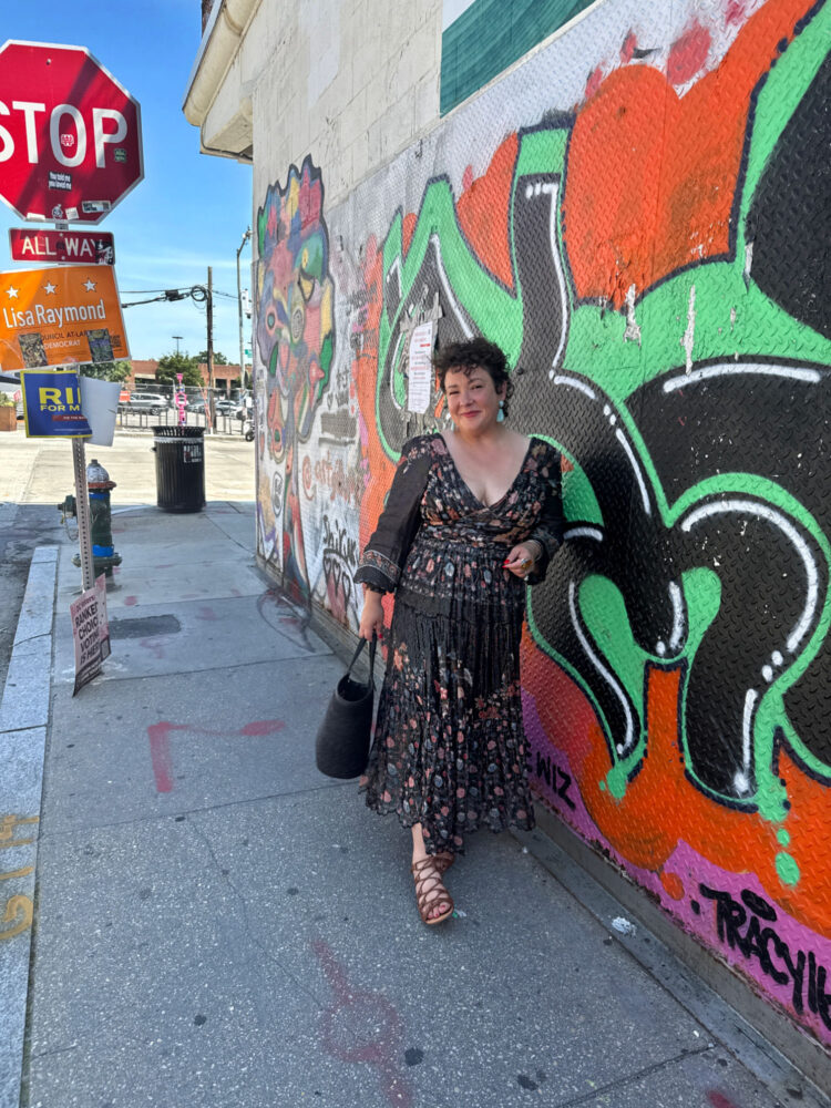 Alison Gary of Wardrobe Oxygen in a black floral Hemant & Nandita boho maxi dress with gold threads, black straw tote, and brown lace-up sandals, leaning against a colorful graffiti wall in Washington DC