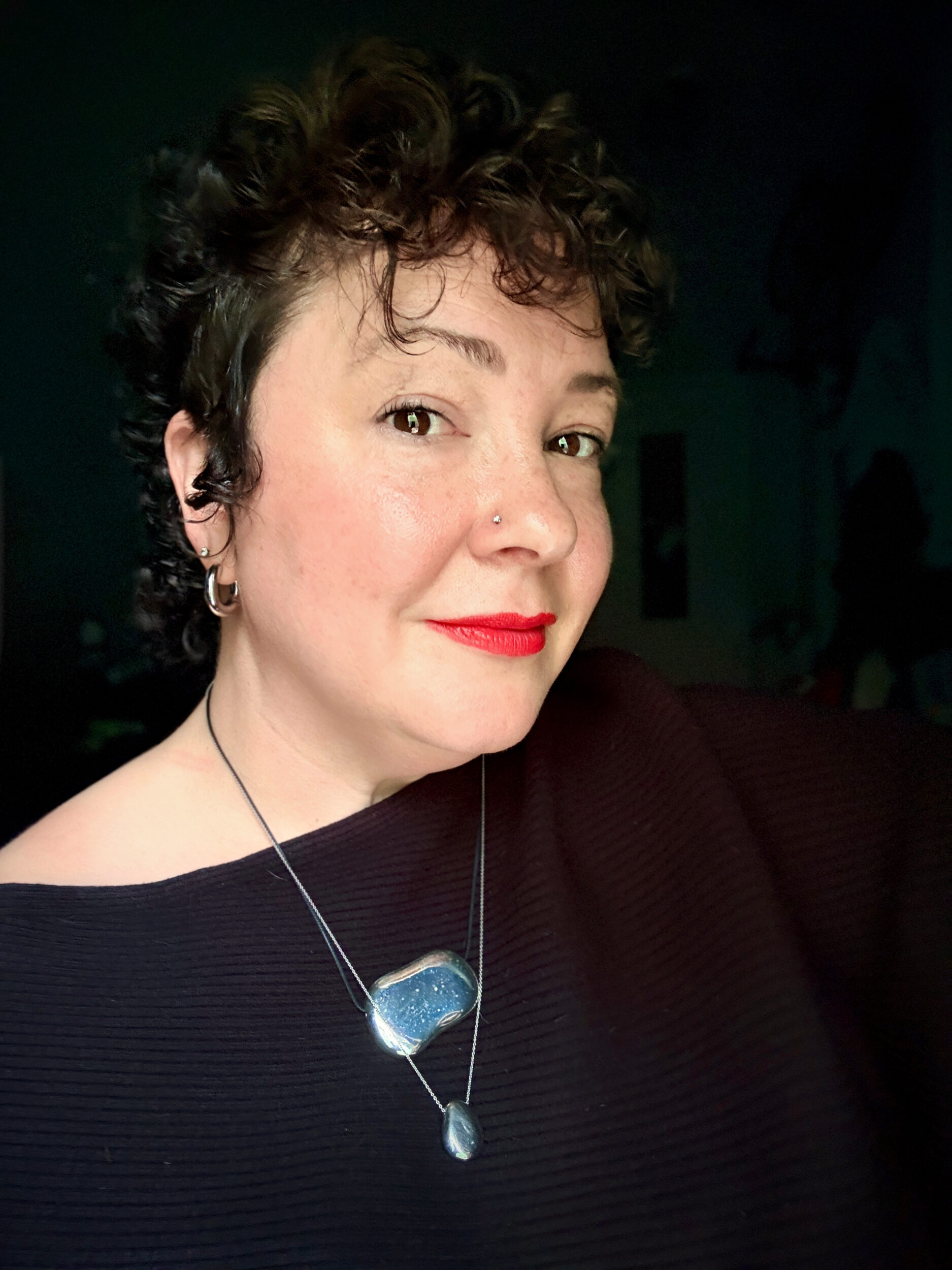 Alison Gary of Wardrobe Oxygen in 2026 wearing a black Reiss Lorna off the shoulder sweater with silver jewelry including the Julia Necklace from Heaven Mayhem. She is not wearing glasses and has on bright red lipstick.